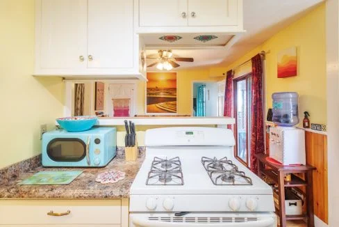 A kitchen with a white gas stove, white cabinets, a microwave oven, a knife block, and a water dispenser. Yellow walls and a ceiling fan in the background.