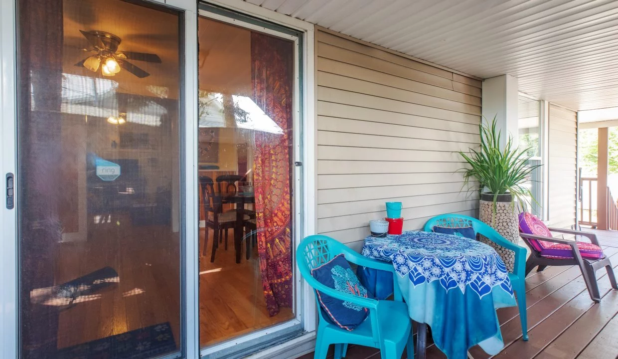 Outdoor patio area with sliding glass door leading inside. Patio includes a small table with a blue-patterned tablecloth and three plastic chairs, one plant, and a lounge chair with a purple cushion.