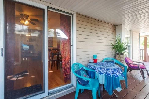 Outdoor patio area with sliding glass door leading inside. Patio includes a small table with a blue-patterned tablecloth and three plastic chairs, one plant, and a lounge chair with a purple cushion.