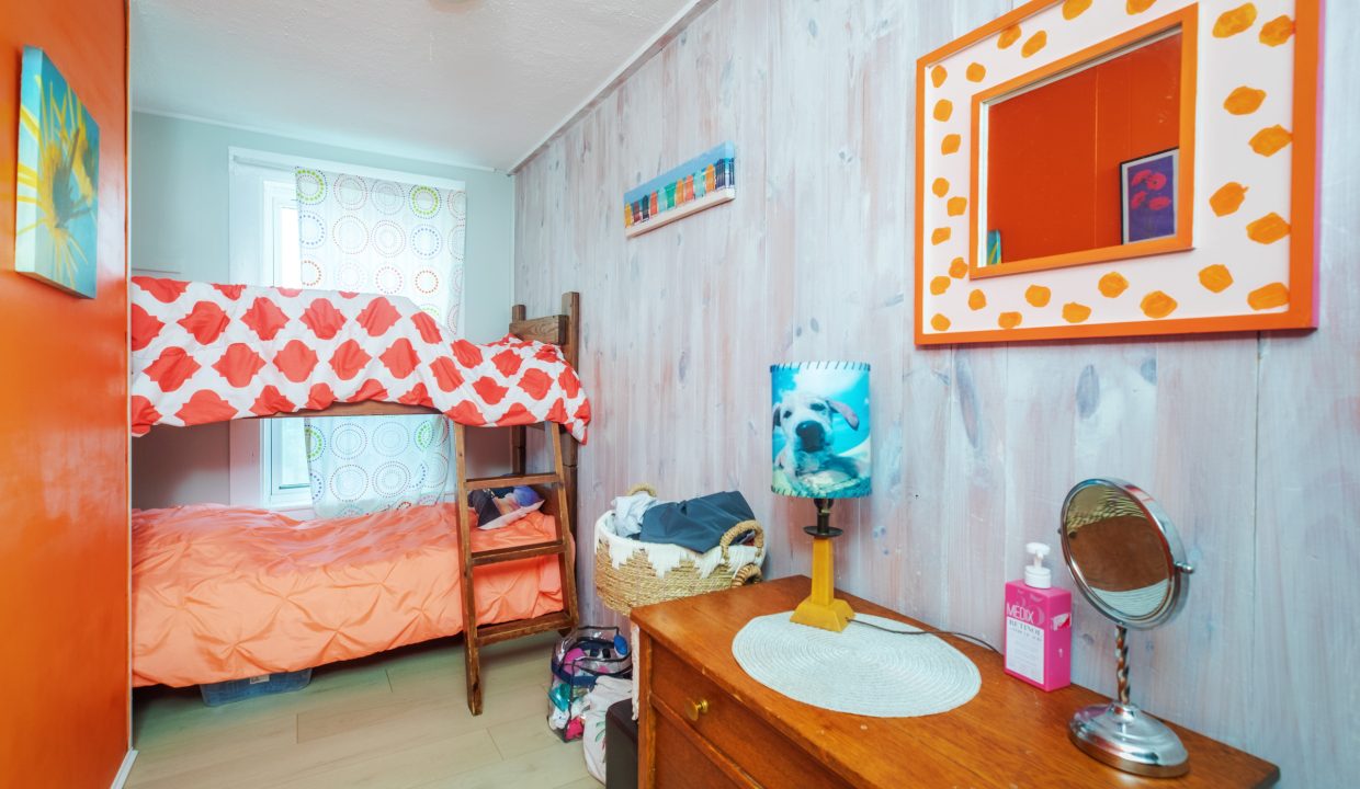 A small bedroom with a wooden bunk bed, orange-accented walls, a mirror on the right wall, a dresser with a round mirror and toiletries, and a window with patterned curtains.