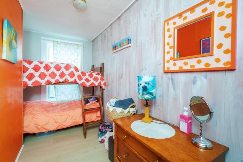 A small bedroom with a wooden bunk bed, orange-accented walls, a mirror on the right wall, a dresser with a round mirror and toiletries, and a window with patterned curtains.