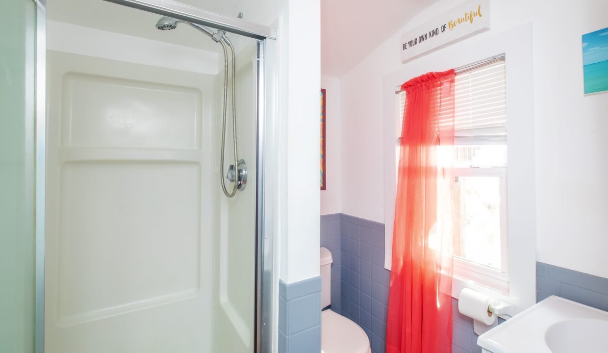 A bathroom with a shower stall, toilet, and sink. Red curtains cover a window, a roll of toilet paper is hanging on the wall, and a colorful sign hangs above the window.