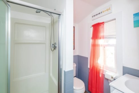 A bathroom with a shower stall, toilet, and sink. Red curtains cover a window, a roll of toilet paper is hanging on the wall, and a colorful sign hangs above the window.