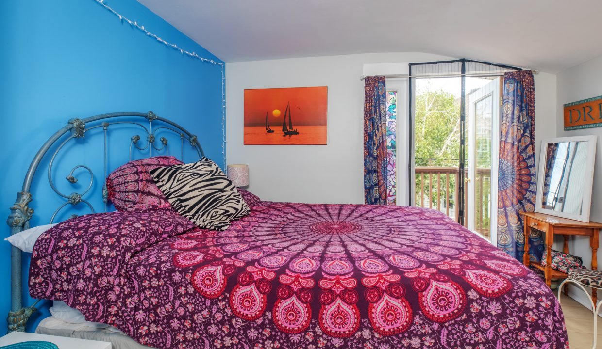 A bedroom with a double bed covered in a purple mandala-patterned duvet, a zebra-print pillow, a metal bed frame, string lights, and a colorful tapestry over the opening to a balcony.