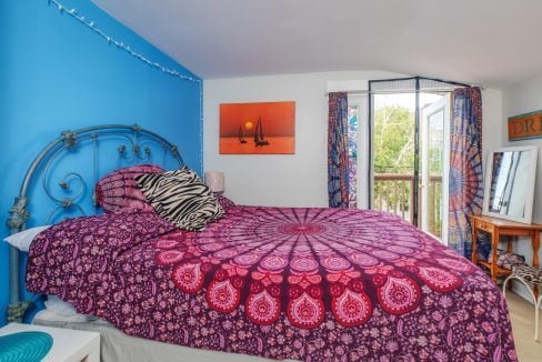 A bedroom with a double bed covered in a purple mandala-patterned duvet, a zebra-print pillow, a metal bed frame, string lights, and a colorful tapestry over the opening to a balcony.