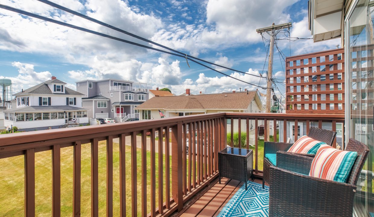 Balcony with wicker furniture, colorful cushions, and a blue geometric rug, overlooking houses, grassy yard, and utility poles against a partly cloudy sky.