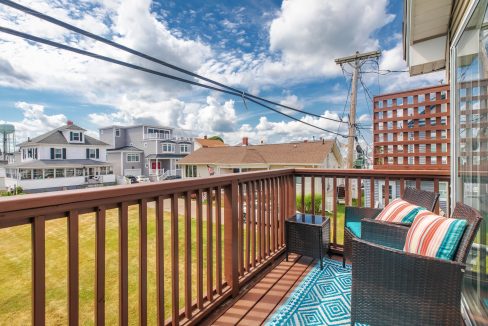 Balcony with wicker furniture, colorful cushions, and a blue geometric rug, overlooking houses, grassy yard, and utility poles against a partly cloudy sky.