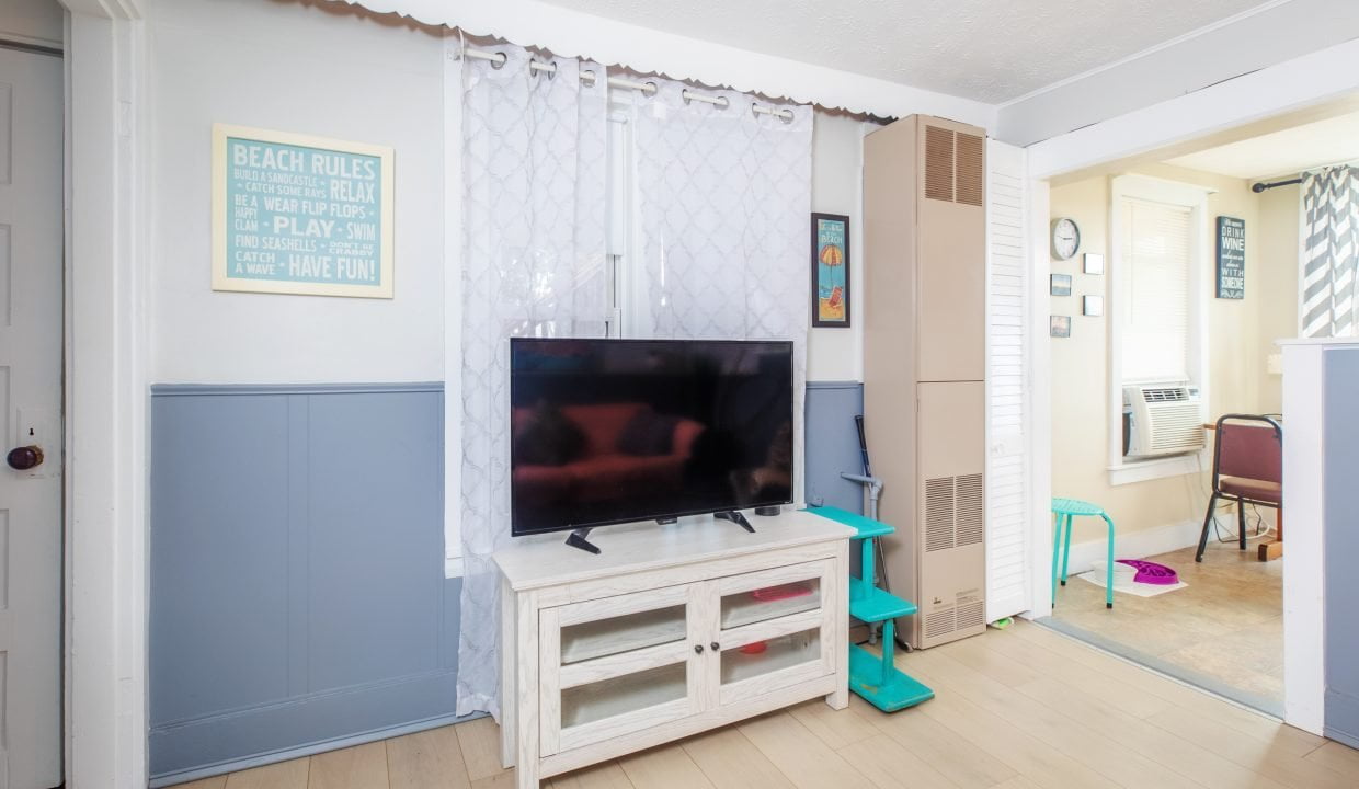 A living room with a flat-screen TV on a white stand, a board with beach rules on the wall, and a tall cardboard box next to green steps. The room extends into a sunny area with an air conditioner.