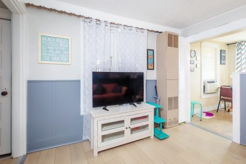 A living room with a flat-screen TV on a white stand, a board with beach rules on the wall, and a tall cardboard box next to green steps. The room extends into a sunny area with an air conditioner.