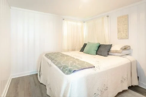 Brightly lit bedroom with a neatly made bed, white walls, and decorative pillows.