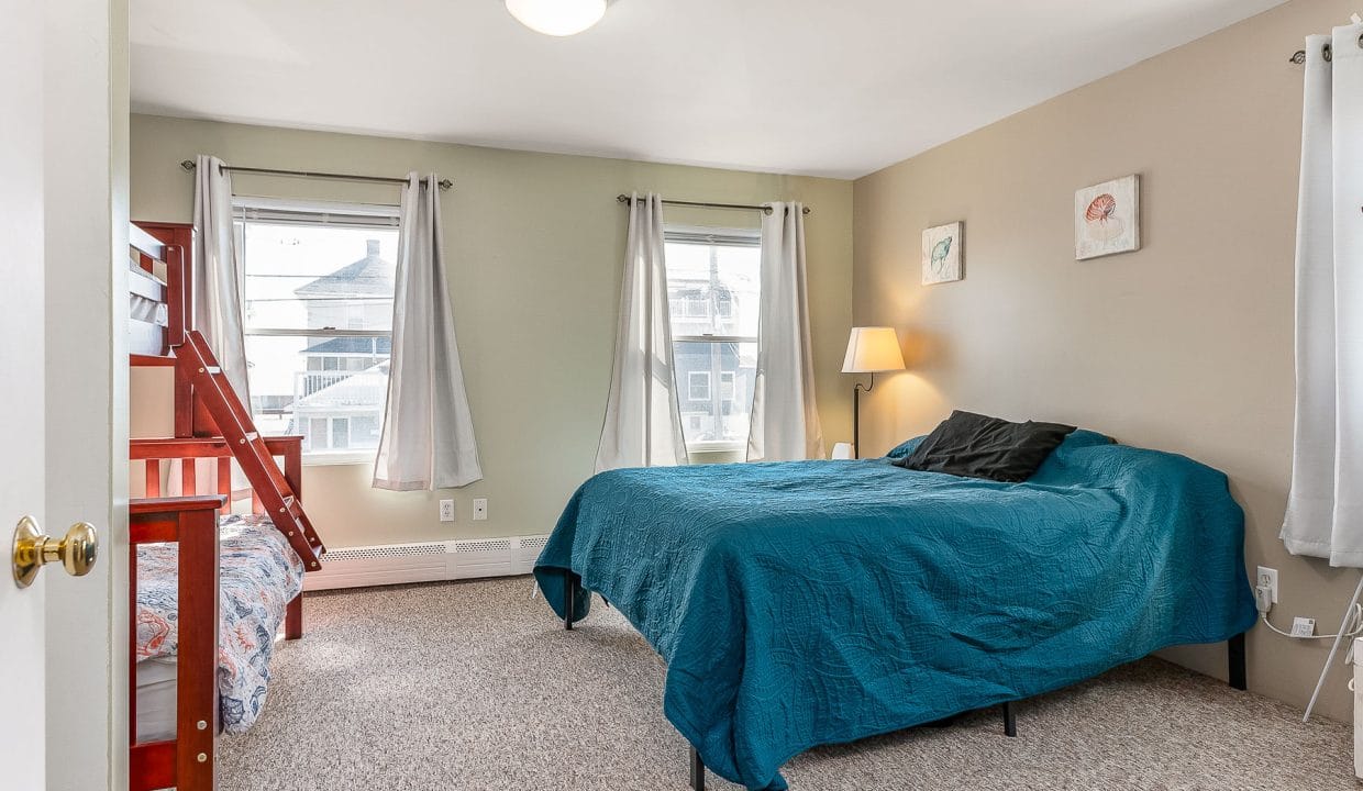 Bedroom with a large bed covered in a teal quilt, a bunk bed with red frame, a lamp, and two framed artworks on the wall. Two windows have sheer curtains. Carpeted floor.