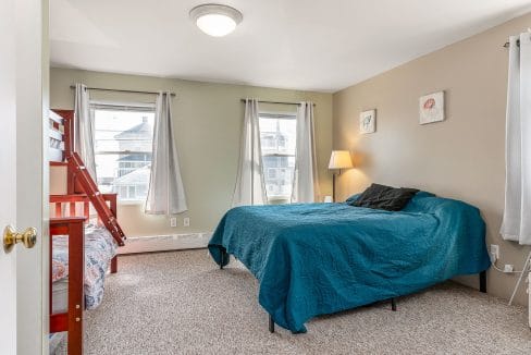 Bedroom with a large bed covered in a teal quilt, a bunk bed with red frame, a lamp, and two framed artworks on the wall. Two windows have sheer curtains. Carpeted floor.