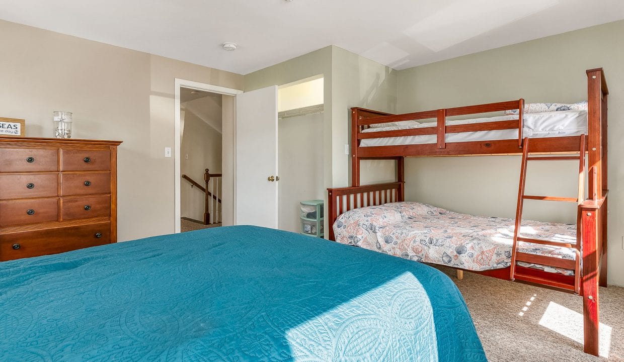 Bedroom with a wooden bunk bed, double bed with blue cover, wooden dresser, and open door leading to a staircase. Natural light illuminates the room.