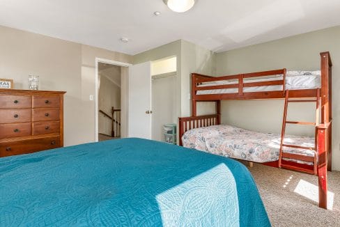 Bedroom with a wooden bunk bed, double bed with blue cover, wooden dresser, and open door leading to a staircase. Natural light illuminates the room.