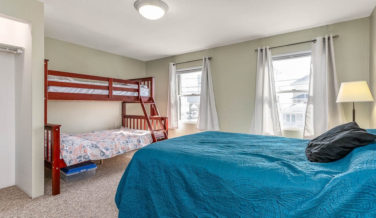 Bedroom with a double bed, a bunk bed, and two windows with white curtains. A lamp stands next to the bed.