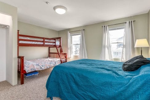 Bedroom with a double bed, a bunk bed, and two windows with white curtains. A lamp stands next to the bed.