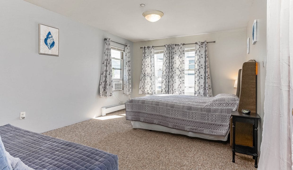 A bedroom with white walls, patterned curtains, a bed, and a blue sofa. A ceiling light is above, and there is artwork on the wall.