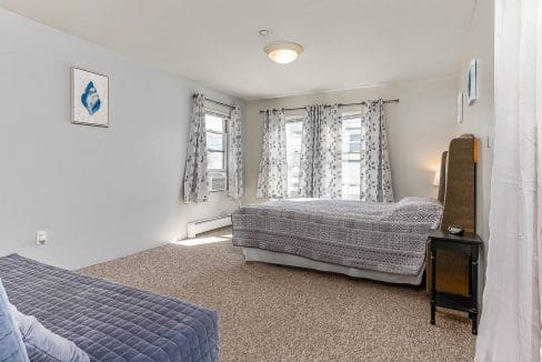 A bedroom with white walls, patterned curtains, a bed, and a blue sofa. A ceiling light is above, and there is artwork on the wall.