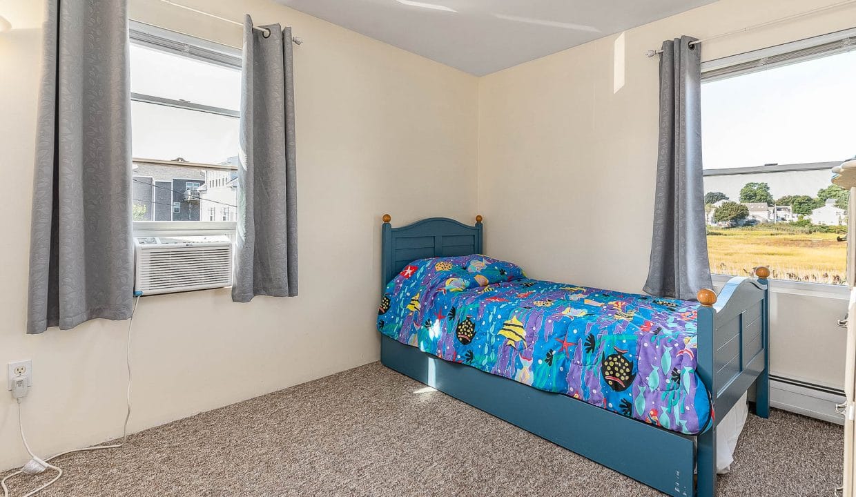 Small bedroom with a blue single bed featuring colorful ocean-themed bedding. Two windows with gray curtains, an air conditioning unit, and beige carpet are also visible.