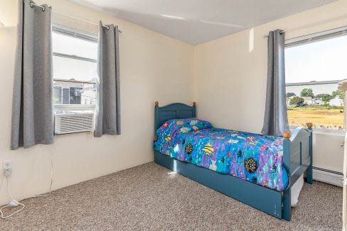 Small bedroom with a blue single bed featuring colorful ocean-themed bedding. Two windows with gray curtains, an air conditioning unit, and beige carpet are also visible.