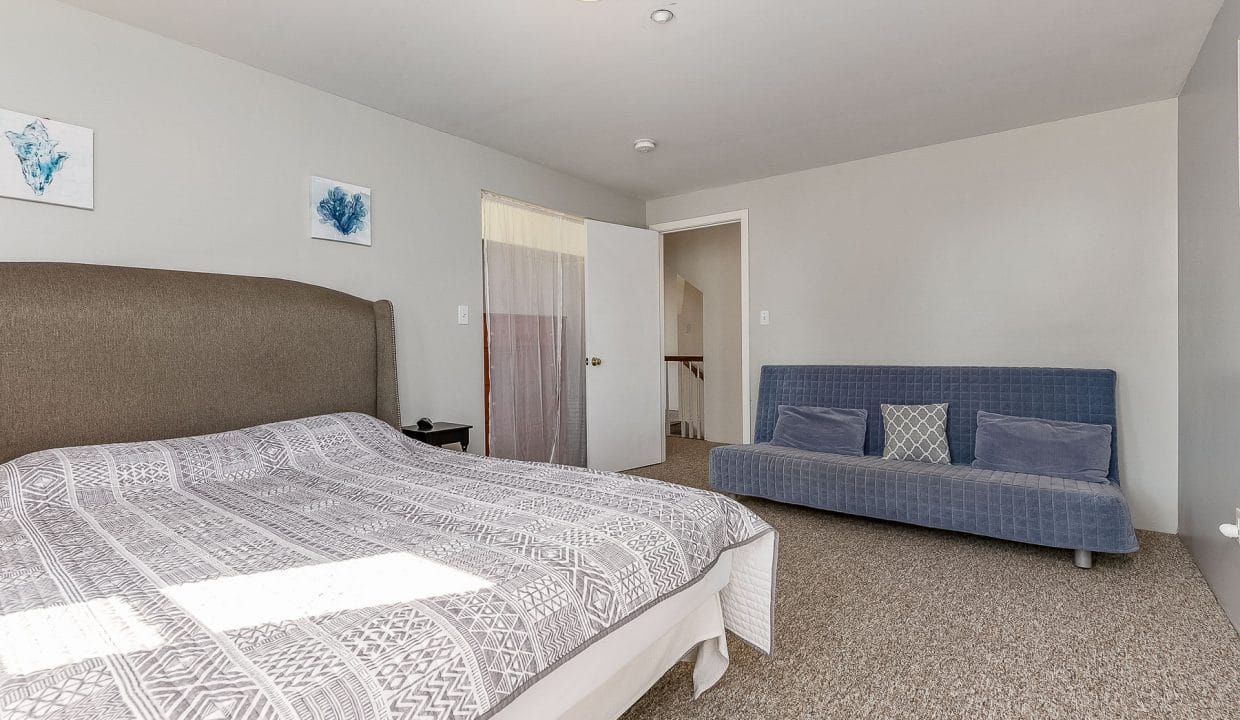 A bedroom with a bed, a gray upholstered headboard, patterned bedspread, and a blue futon. Two abstract artworks hang on the wall, and a doorway leads to a hallway with a staircase.