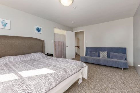 A bedroom with a bed, a gray upholstered headboard, patterned bedspread, and a blue futon. Two abstract artworks hang on the wall, and a doorway leads to a hallway with a staircase.