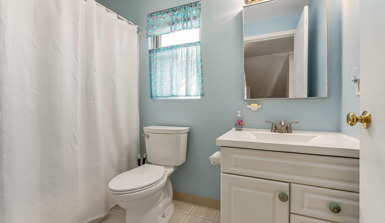 A clean bathroom with a toilet, sink with cabinet, mirror, and shower curtain. The walls are light blue, and there is a window with a patterned curtain.