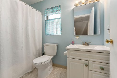 A clean bathroom with a toilet, sink with cabinet, mirror, and shower curtain. The walls are light blue, and there is a window with a patterned curtain.