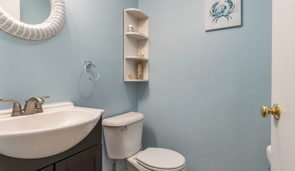 A small bathroom with blue walls features a white sink and toilet, circular mirror, corner shelf, and crab-themed wall art.