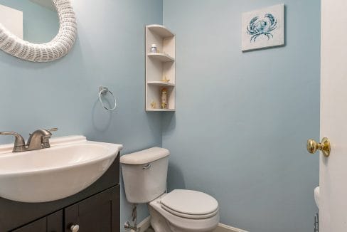 A small bathroom with blue walls features a white sink and toilet, circular mirror, corner shelf, and crab-themed wall art.