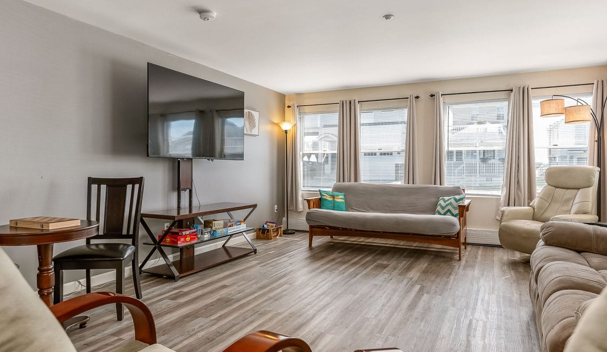 A living room with a wall-mounted TV, a futon, two armchairs, a small table, and large windows with curtains.