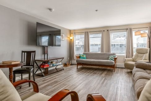 A living room with a wall-mounted TV, a futon, two armchairs, a small table, and large windows with curtains.