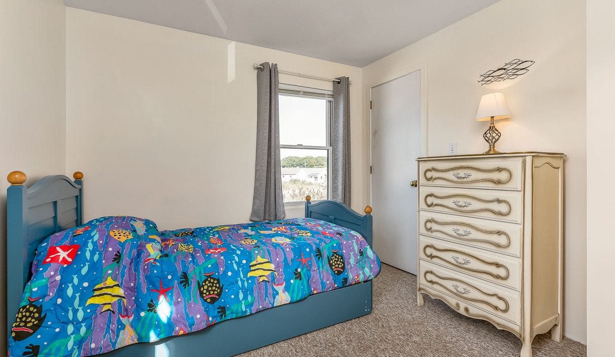 A small bedroom with a blue bed featuring an ocean-themed comforter, a white dresser, lamp, gray curtains, and a window with a view.