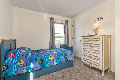 A small bedroom with a blue bed featuring an ocean-themed comforter, a white dresser, lamp, gray curtains, and a window with a view.