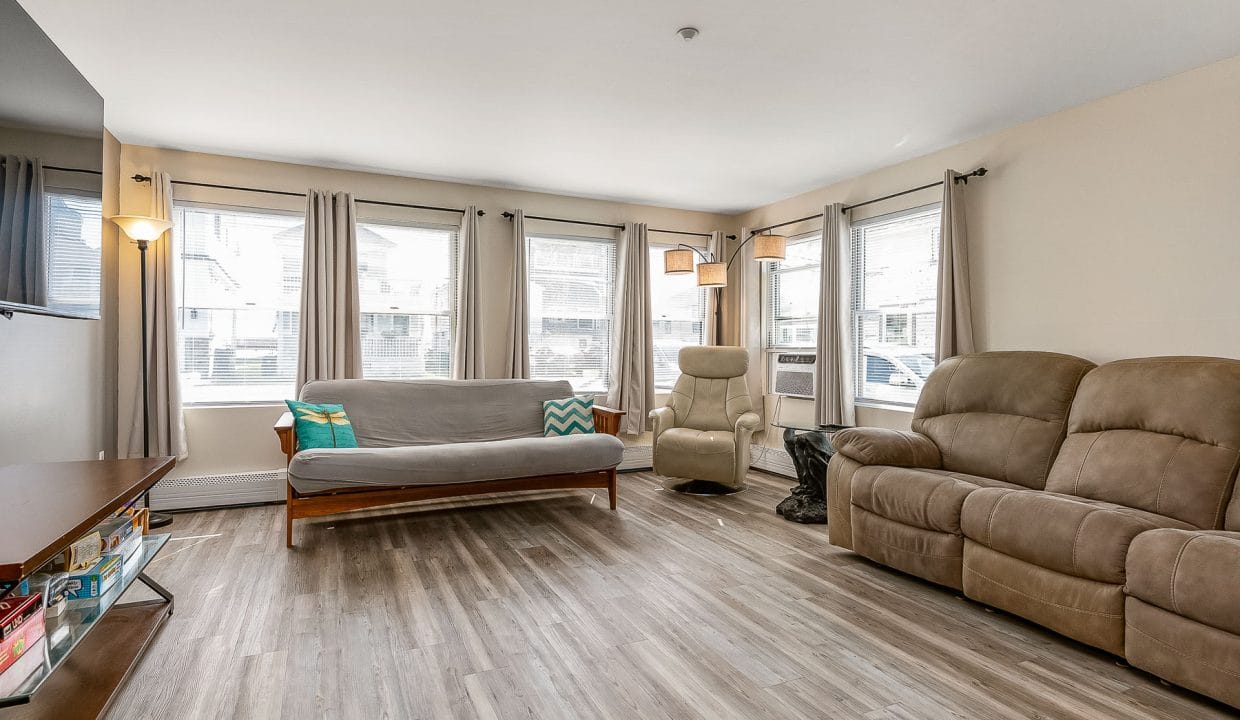 A living room with a wooden floor, featuring a sofa, reclining chair, and loveseat, natural light from multiple windows, and a TV stand with books.