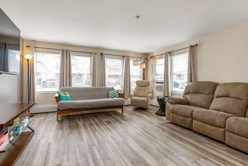 A living room with a wooden floor, featuring a sofa, reclining chair, and loveseat, natural light from multiple windows, and a TV stand with books.