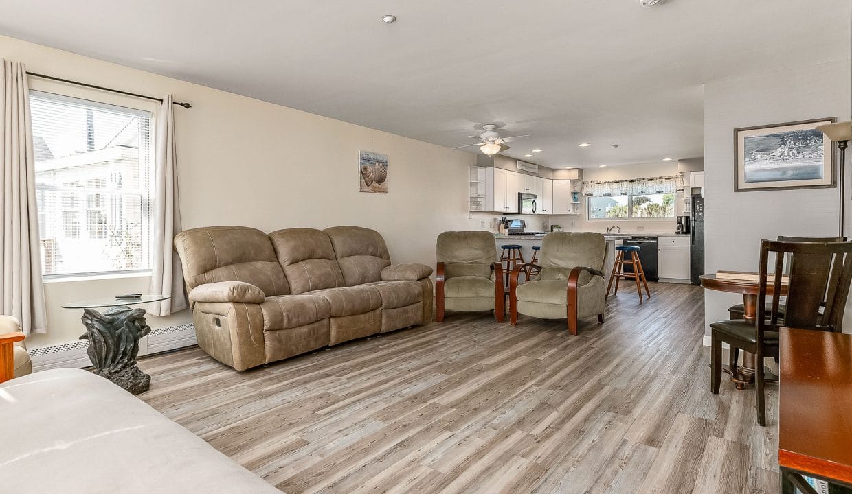 Spacious living room with beige sofas, wooden chairs, and dining table. Open kitchen with white cabinets in the background. Large window with curtains on the left. Hardwood floor throughout.