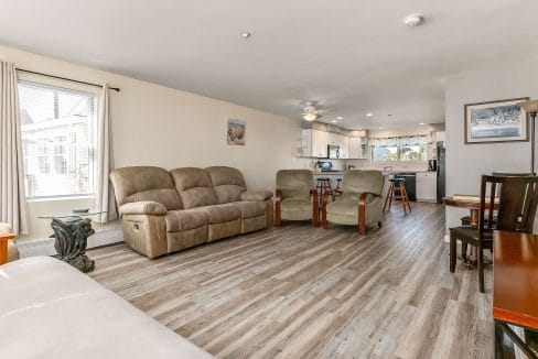 Spacious living room with beige sofas, wooden chairs, and dining table. Open kitchen with white cabinets in the background. Large window with curtains on the left. Hardwood floor throughout.