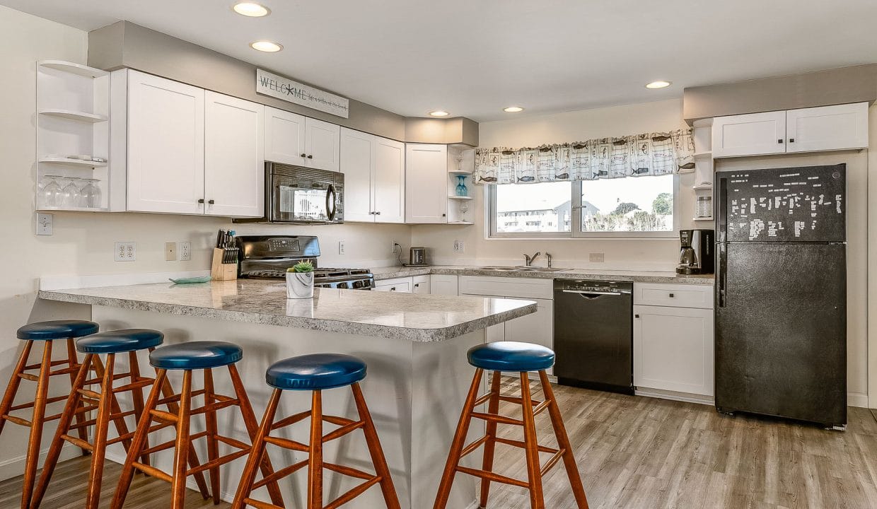 Modern kitchen with white cabinets, a black fridge, stainless steel appliances, and an island with four wooden stools. The room has wood flooring and a window with a patterned curtain.