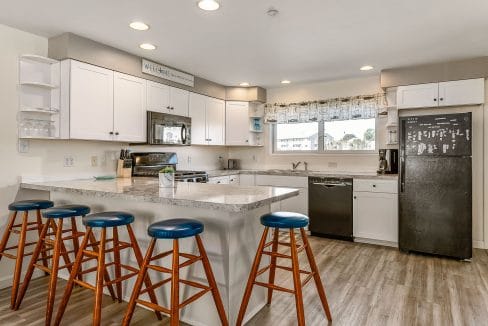 Modern kitchen with white cabinets, a black fridge, stainless steel appliances, and an island with four wooden stools. The room has wood flooring and a window with a patterned curtain.