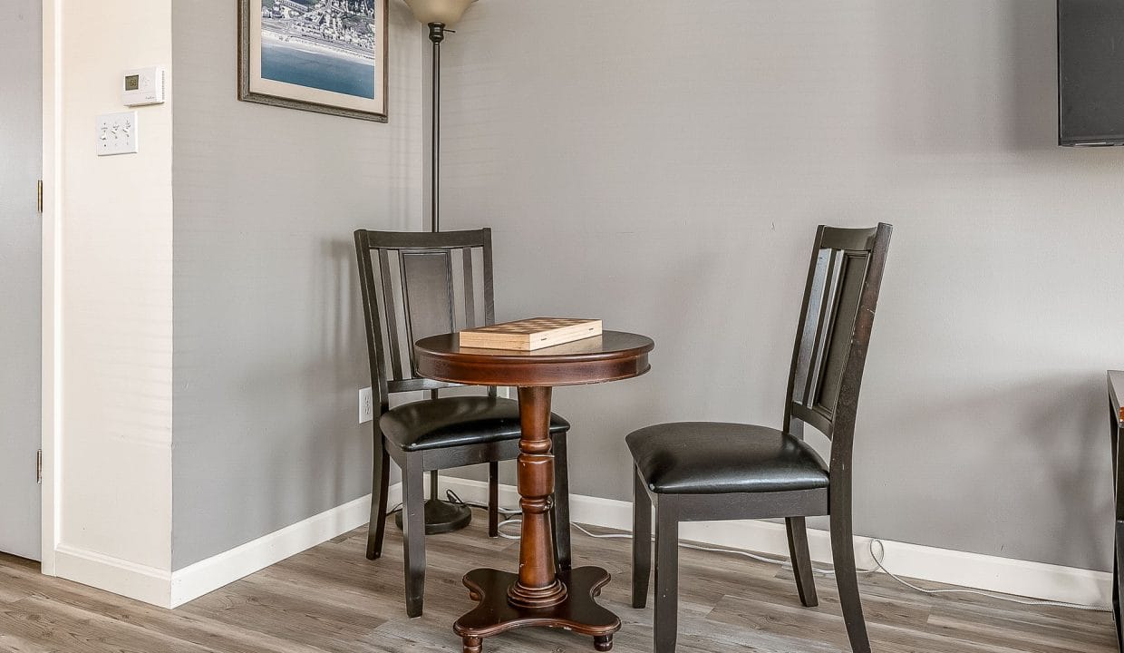 A small round table with two black chairs is placed in a corner of a room with gray walls. A floor lamp and a framed picture are on the walls. A wooden game board is on the table.