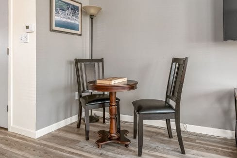 A small round table with two black chairs is placed in a corner of a room with gray walls. A floor lamp and a framed picture are on the walls. A wooden game board is on the table.