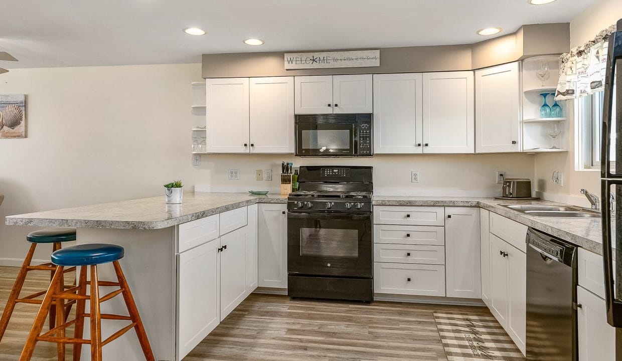 A modern kitchen with white cabinets, black appliances, and a light gray countertop. Two wooden stools with blue seats are placed by the counter. Bright overhead lighting illuminates the space.