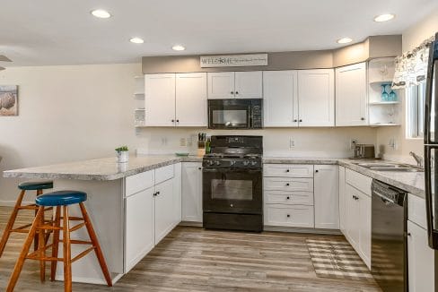 A modern kitchen with white cabinets, black appliances, and a light gray countertop. Two wooden stools with blue seats are placed by the counter. Bright overhead lighting illuminates the space.