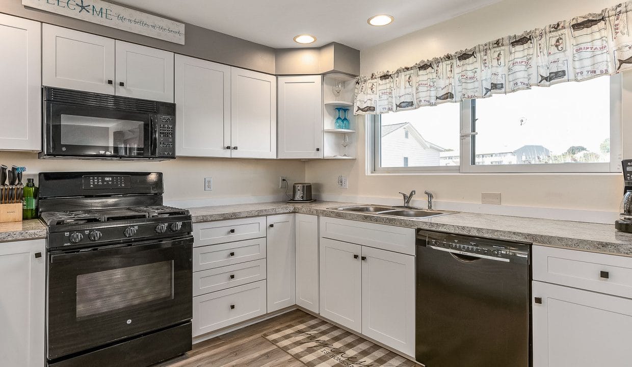 Modern kitchen with white cabinets, black appliances, and wood flooring. A welcome sign is above the cabinets, and there’s a large window over the sink.