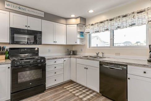 Modern kitchen with white cabinets, black appliances, and wood flooring. A welcome sign is above the cabinets, and there’s a large window over the sink.