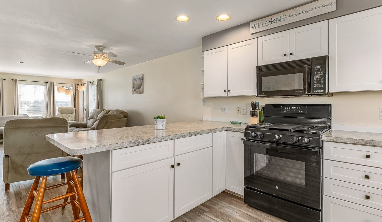 Modern kitchen with white cabinets, gray countertops, black appliances, and light wood flooring. A bar stool is at the counter, and an adjoining living area is visible in the background.