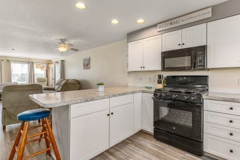 Modern kitchen with white cabinets, gray countertops, black appliances, and light wood flooring. A bar stool is at the counter, and an adjoining living area is visible in the background.