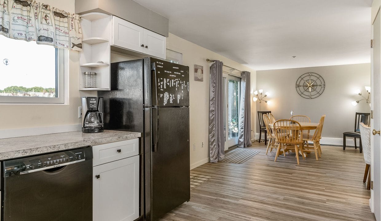 Kitchen with black appliances and white cabinets, opening to a dining area with a wooden table and chairs. Neutral-toned walls and a floor lamp are visible.