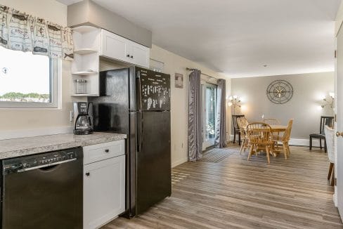 Kitchen with black appliances and white cabinets, opening to a dining area with a wooden table and chairs. Neutral-toned walls and a floor lamp are visible.